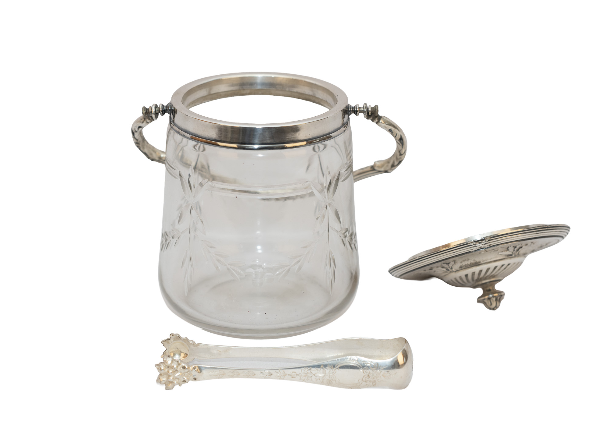 Seau à biscuits en verre taillé ouvert, avec anse rabattue vers l’arrière, couvercle décoré et pince à sucre au premier plan.